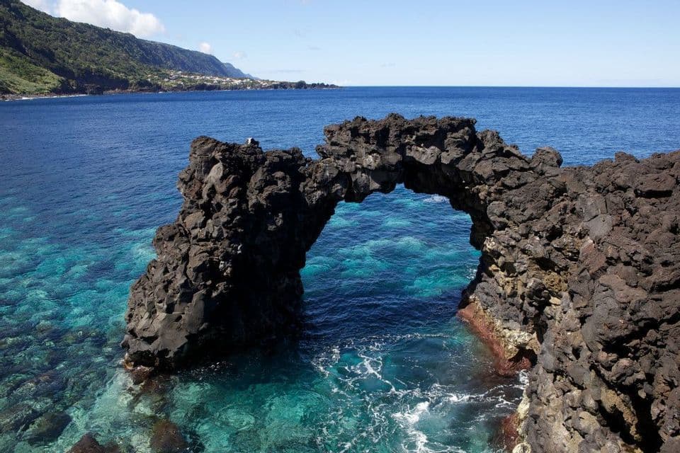 Una formazione ad arco di roccia vulcanica si erge sull'oceano turchese con una costa verde e collinare sullo sfondo.