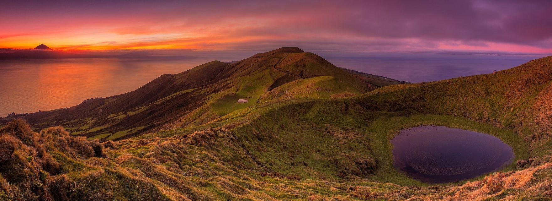 Vista panoramica di una costa verdeggiante e collinare con un laghetto che si affaccia sull'oceano e un vulcano lontano al tramonto.