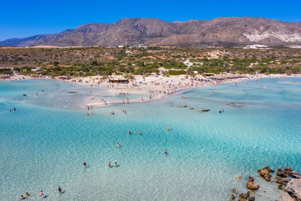 Vista aerea di una spiaggia affollata con persone che nuotano in acque basse e turchesi, con montagne aride sullo sfondo.