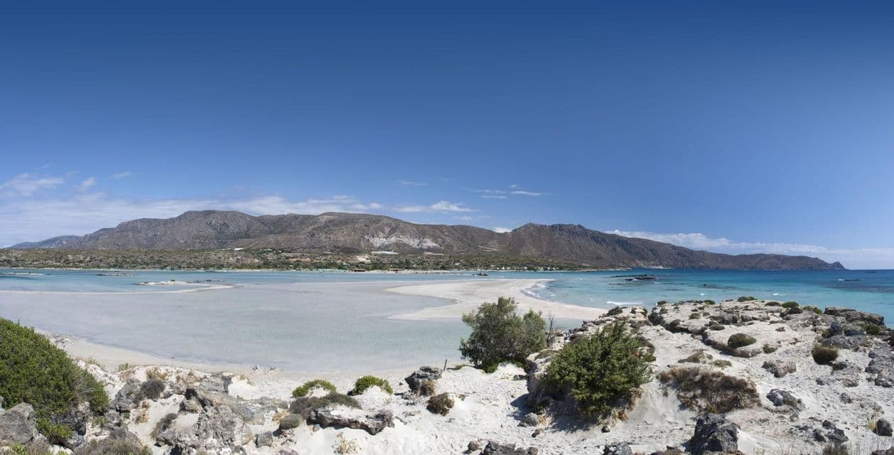 Una vista panoramica di una laguna turchese poco profonda e di una spiaggia di sabbia bianca, con montagne aride sullo sfondo sotto un cielo azzurro.