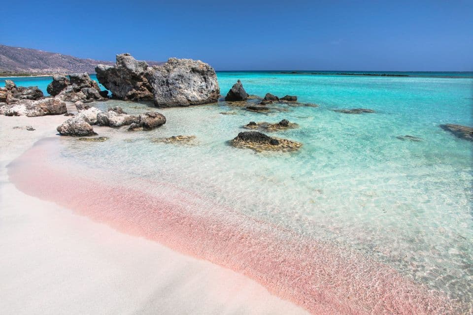 Una costa con sabbia rosa incontra acque limpide e turchesi punteggiate da grandi rocce sotto un cielo azzurro e soleggiato.