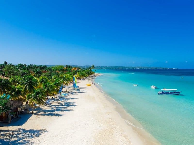 Blick von oben auf einen tropischen weißen Sandstrand mit Palmen und Booten im türkisfarbenen Wasser unter klarem blauem Himmel.
