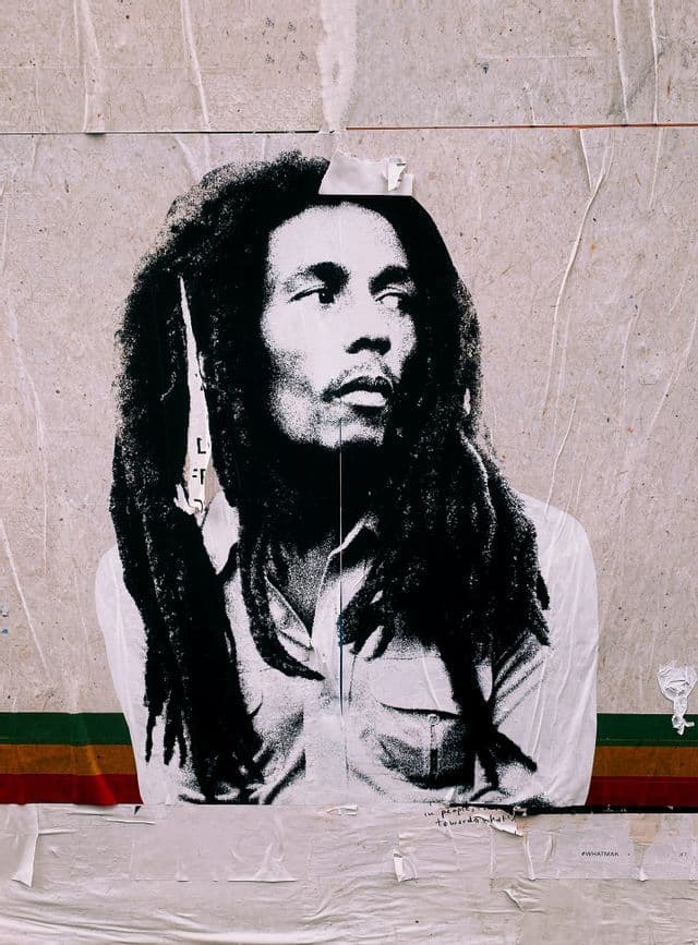 Ein Schwarz-Weiß-Paste-up-Plakat von Bob Marley mit Dreadlocks löst sich von einer strukturierten Wand über einem rot-gelb-grünen Streifen.