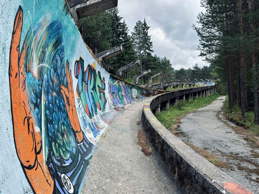 Una pista da bob in cemento abbandonata, coperta di graffiti colorati, si snoda attraverso una foresta di alti pini.