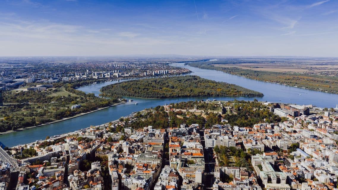 Una vista aerea di una vasta città con un ampio fiume che circonda una grande isola boscosa sotto un cielo azzurro.