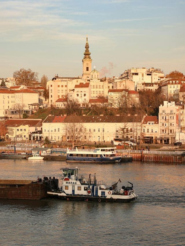 Un rimorchiatore spinge una chiatta lungo un fiume di fronte a uno skyline cittadino con un alto campanile sotto un cielo fosco.