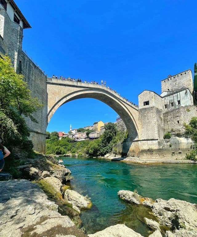 Un grande ponte ad arco in pietra con persone sopra attraversa un fiume turchese, con una città sulla collina sotto un cielo azzurro.