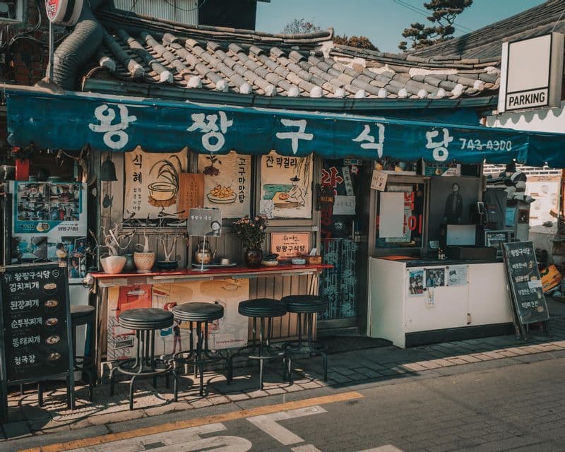 The storefront of a traditional Korean food stall with a blue awning, a red counter, and empty stools on a sunny street.