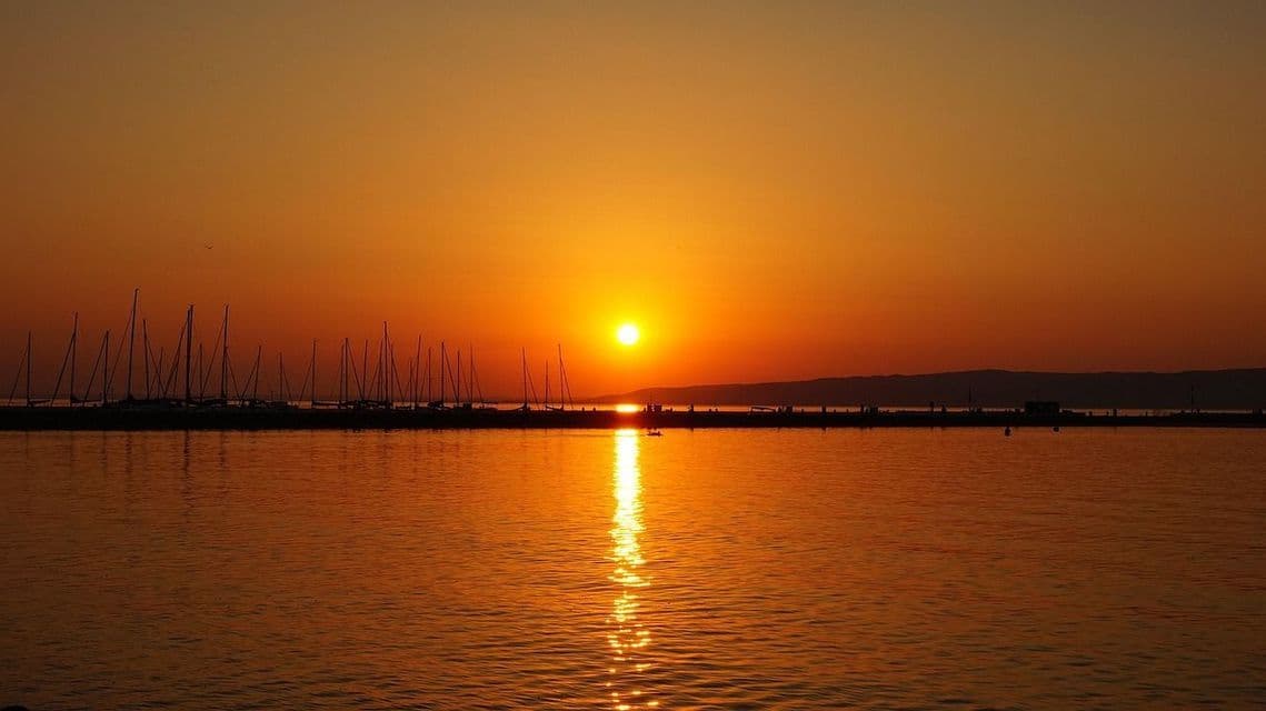 Un sole arancione tramonta su uno specchio d'acqua calmo, stagliando un porto pieno di barche a vela contro l'orizzonte.