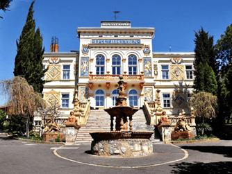 Un edificio ornato, color crema, con una grande scalinata si erge dietro una fontana in pietra, affiancato da alberi sotto un cielo azzurro e limpido.