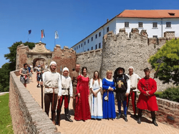 Un gruppo di persone in costume storico posa per una foto su un ponte di mattoni davanti a un castello di pietra.