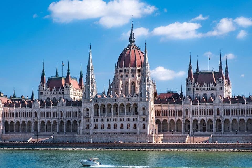 Un motoscafo costeggia un grande e ornato edificio neogotico sulla riva di un fiume sotto un cielo blu con nuvole bianche.