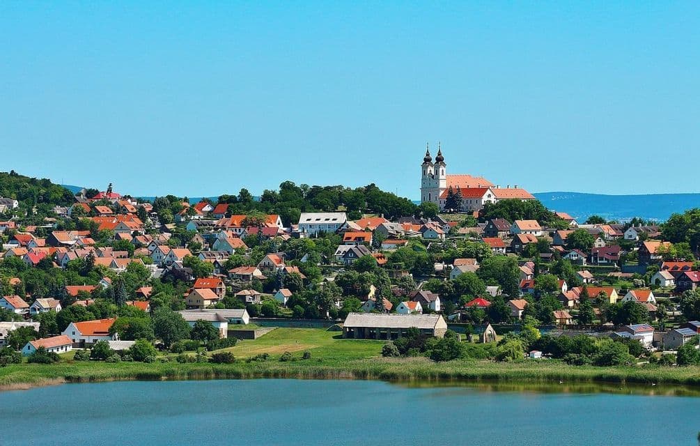 Un borgo con case dai tetti rossi e una chiesa a due torri su una verde collina, visibile da oltre un lago sotto un cielo blu chiaro.