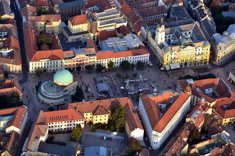 Una veduta aerea di una piazza storica affollata di persone, circondata da edifici con tetti in tegole rosse e una grande cupola verde.