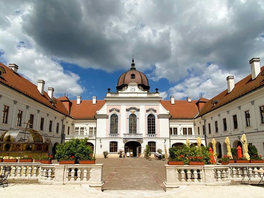Un palazzo simmetrico bianco e rosa con un centro a cupola e tetti rossi si erge in un cortile pavimentato sotto un cielo drammatico e nuvoloso.
