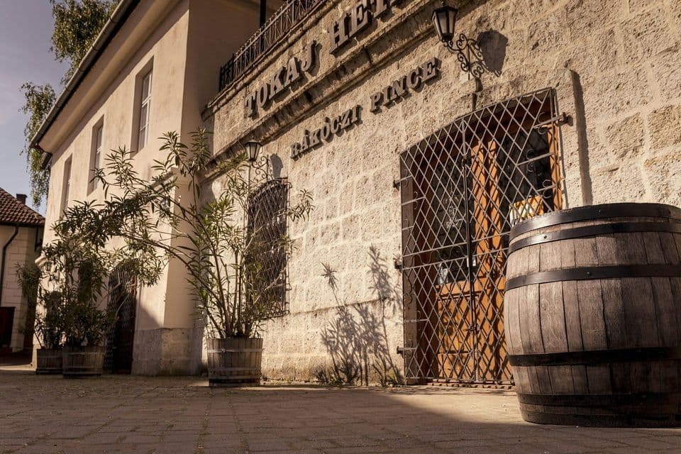 L'ingresso di un edificio in pietra con una grande botte di legno e piante in vaso su un cortile lastricato sotto la luce del sole.