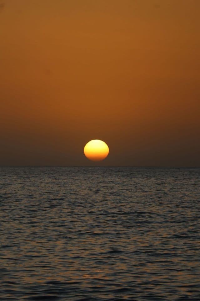 Eine große gelbe Sonne hängt knapp über dem dunklen, welligen Ozean unter einem tief orangefarbenen Himmel.