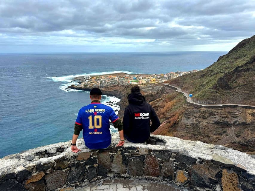 Ein WeRoad Gruppenleiter und eine weitere Person sitzen auf einer Steinmauer an einer Klippe und blicken auf eine Küstenstadt und das Meer.