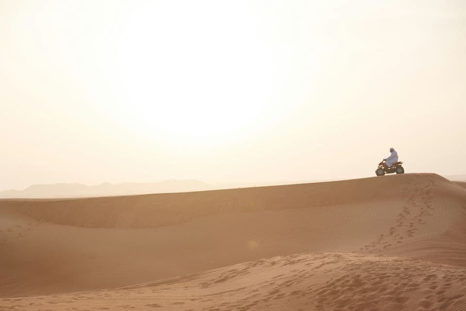 A person wearing white robes rides a quad bike on the crest of a large desert sand dune under a hazy sky.