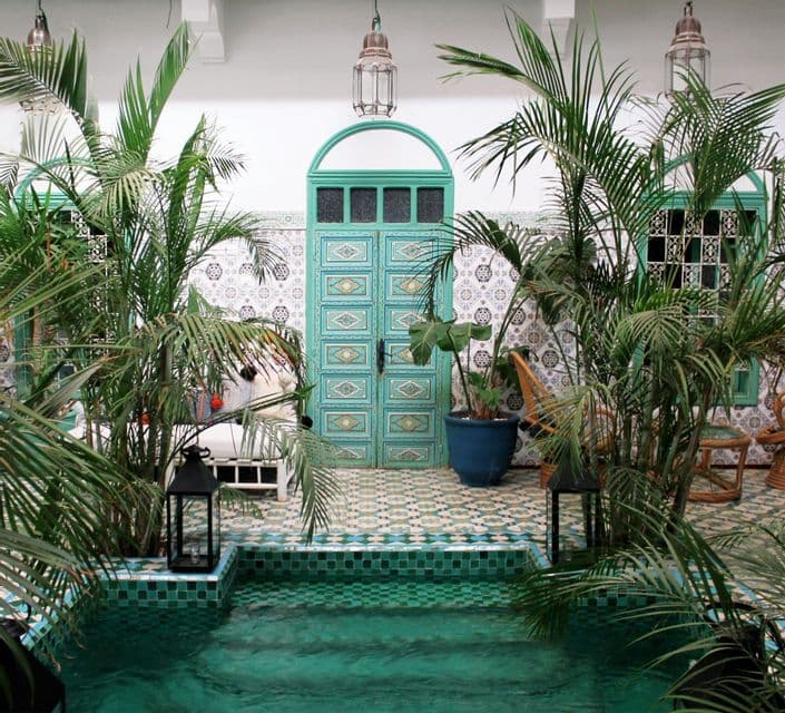 A small pool with green tiles sits in a courtyard with palm plants, patterned tiles, and an ornate turquoise door.