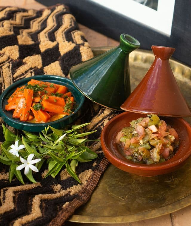 Two small tagines with cooked carrots and a vegetable salad sit on a brass tray next to jasmine flowers.