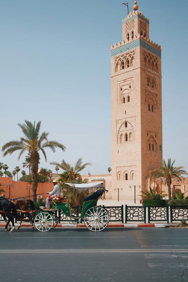 A green horse-drawn carriage travels down a street in front of a tall stone minaret, with palm trees under a clear sky.