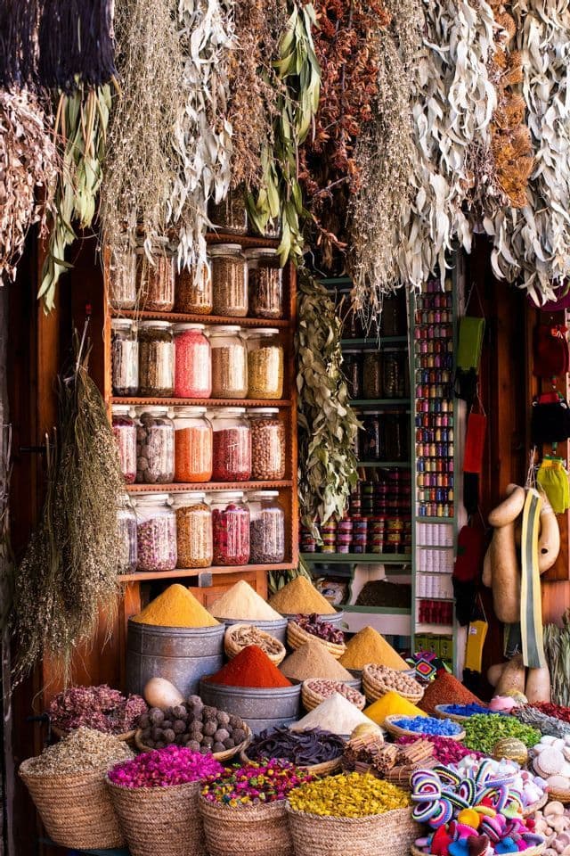 Colorful spices, dried herbs, and flowers displayed in baskets and jars at a busy market stall with more herbs hanging from above.