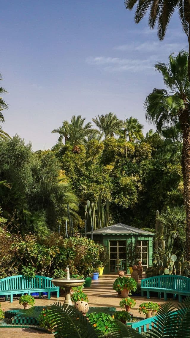 A lush garden courtyard with turquoise benches, a small fountain, and a green pavilion surrounded by palm trees and cacti.