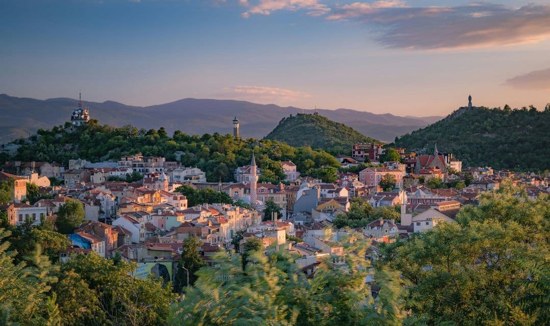 Un paysage urbain de bâtiments colorés nichés parmi des collines verdoyantes, avec plusieurs monuments visibles sous un ciel de coucher de soleil.