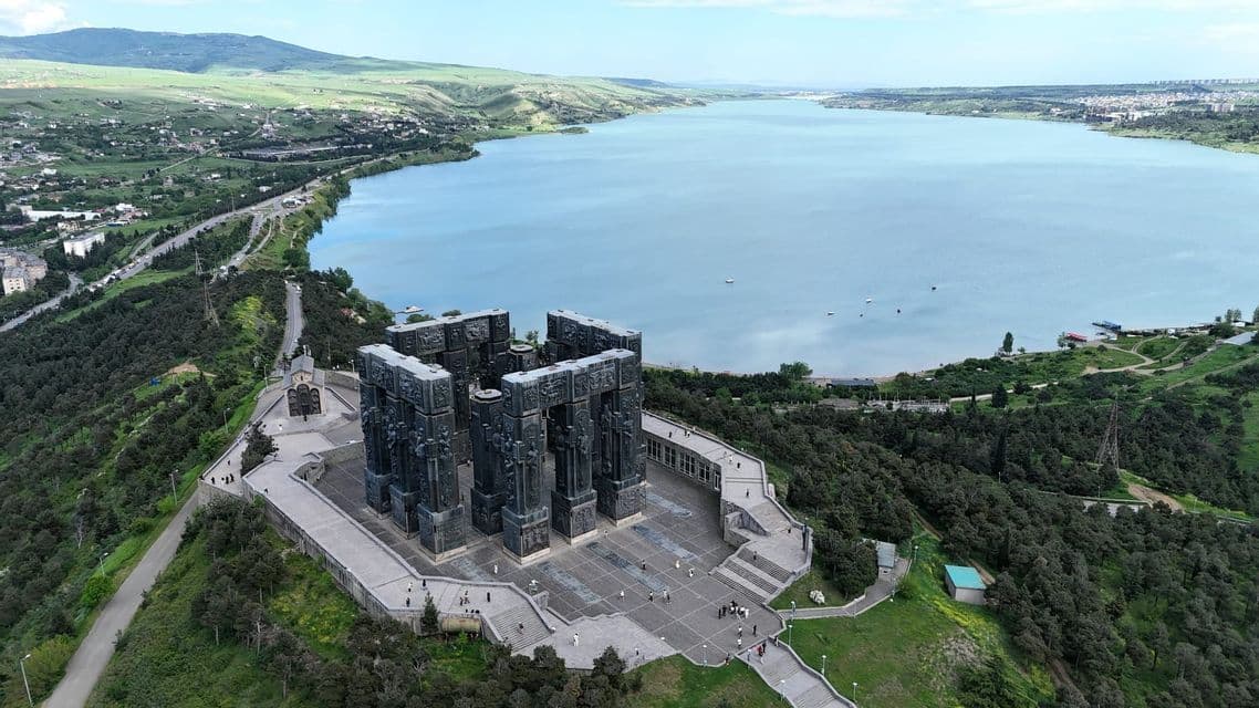 Vue aérienne d'un grand monument sombre aux piliers sculptés, situé sur une esplanade en hauteur, surplombant un vaste lac bleu et des collines verdoyantes.
