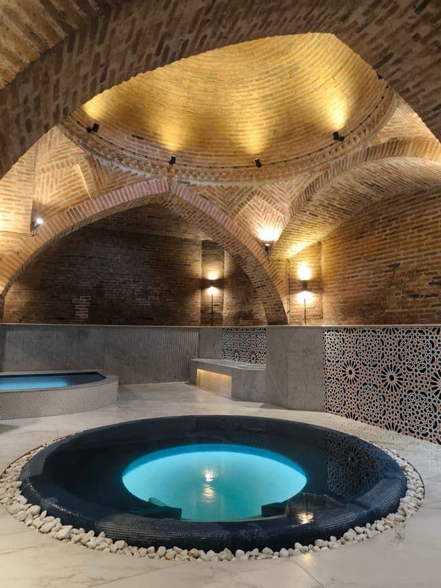 Deux piscines éclairées se trouvent sur un sol en marbre, à l'intérieur d'un spa aux plafonds voûtés en briques et aux murs ornés de carrelage.