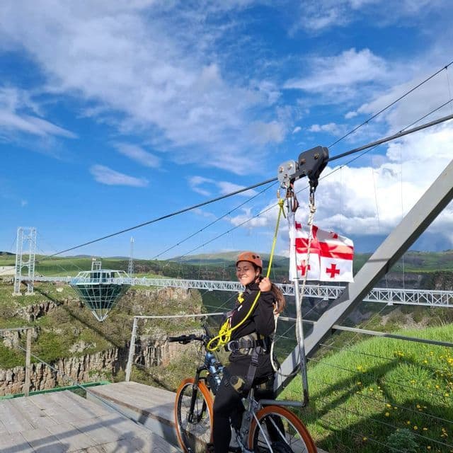 Une femme souriante en casque assise sur un vélo attaché à une tyrolienne au-dessus d'un canyon, avec un grand pont de verre en arrière-plan.