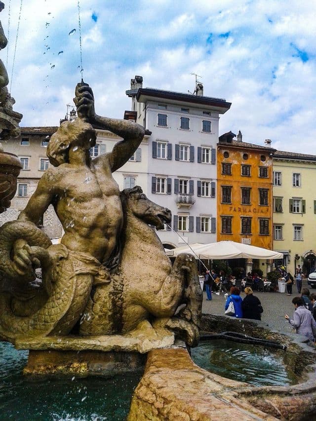Una fontana in pietra con scultura di tritone che spruzza acqua in una piazza europea con edifici colorati.