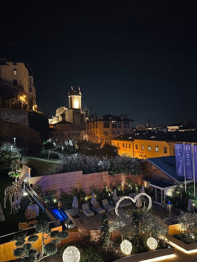 Una vista notturna di una terrazza d'albergo illuminata con statue di giraffa, che si affaccia su una città collinare con un antico castello.