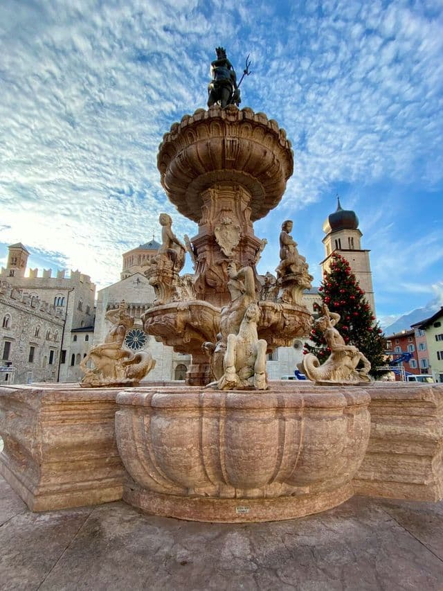 Una fontana in pietra ornata con una statua di Nettuno si erge in una piazza cittadina, con un albero di Natale e edifici storici sullo sfondo.