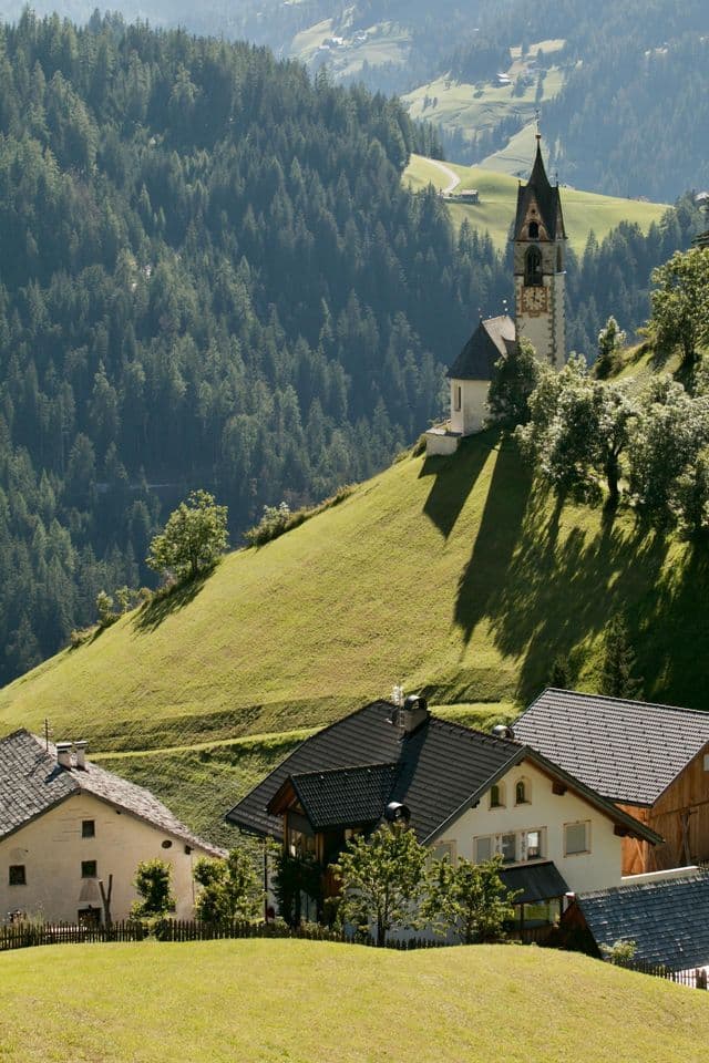 Una chiesa bianca con un'alta torre campanaria sorge su una ripida collina erbosa sopra un villaggio, con una fitta pineta sullo sfondo.