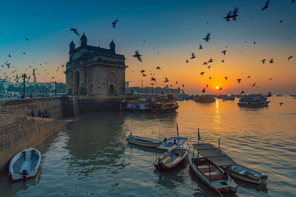 Un arco de piedra domina un puerto con barcos al amanecer, mientras una bandada de pájaros vuela por el cielo colorido.