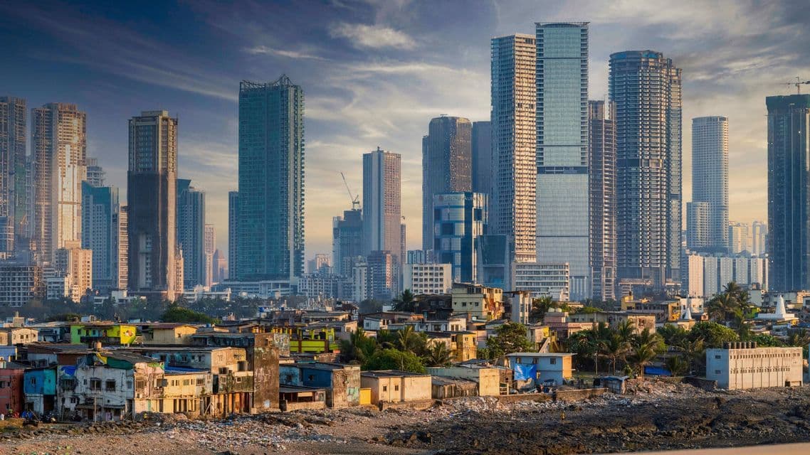 Modernos rascacielos se elevan sobre un primer plano de edificios bajos y desgastados, y una costa rocosa bajo un cielo nublado.