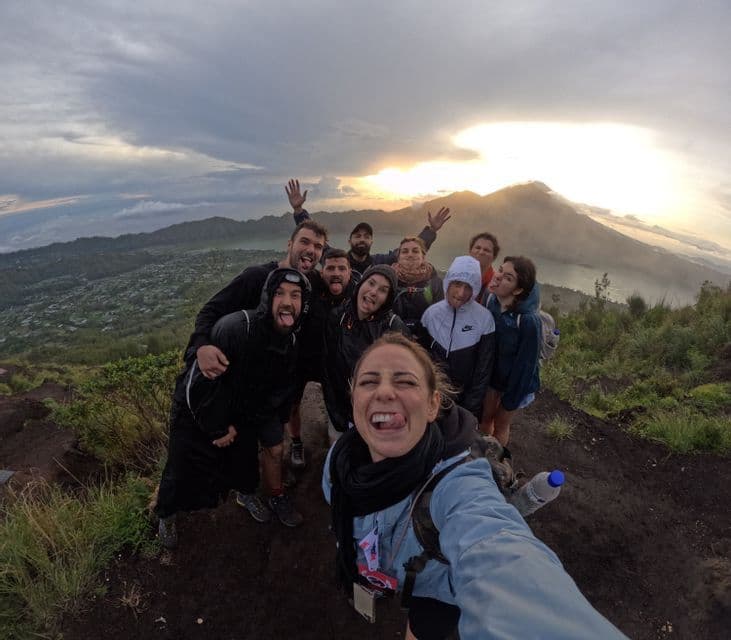 Un viaje en grupo de WeRoad se toma una selfie divertida con ojo de pez en la cima de una montaña al amanecer, con vistas a un volcán y un lago.