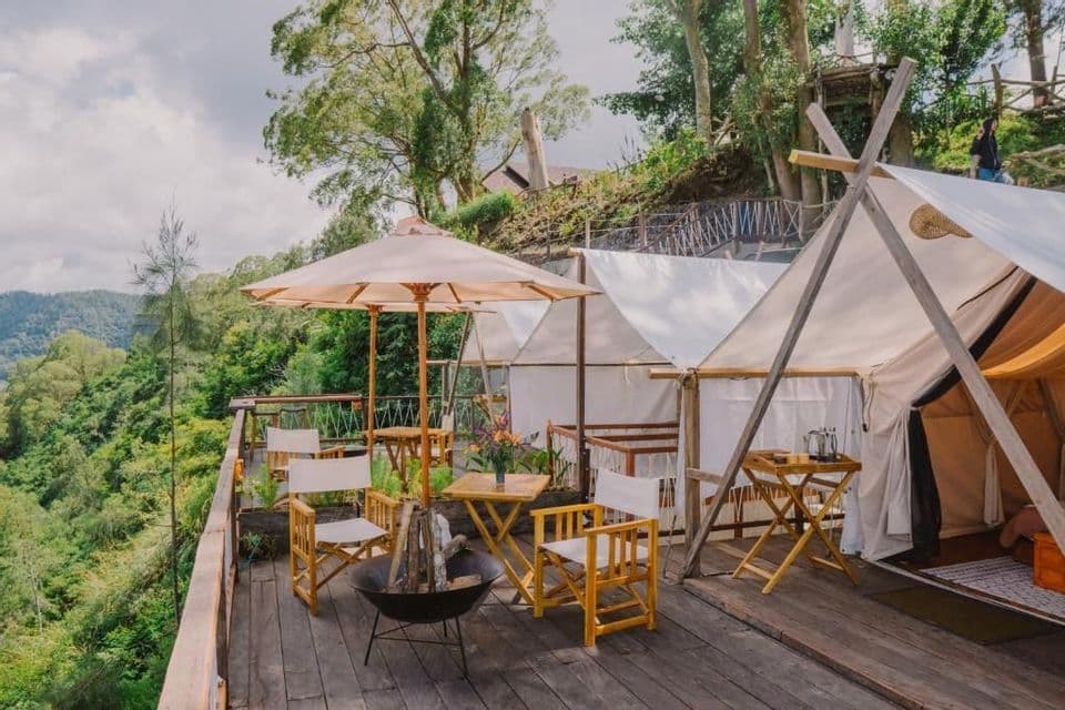 Ein Glamping-Zeltplatz auf einer Holzterrasse mit weißen Zelten, Stühlen, einem Sonnenschirm und einer Feuerstelle mit Blick auf einen üppigen Wald.