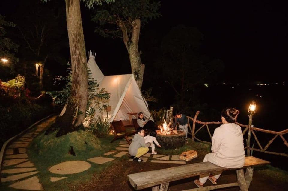 Eine Gruppe von Menschen versammelt sich nachts um ein Lagerfeuer neben einem beleuchteten Tipi-Zelt in einem Waldgebiet.