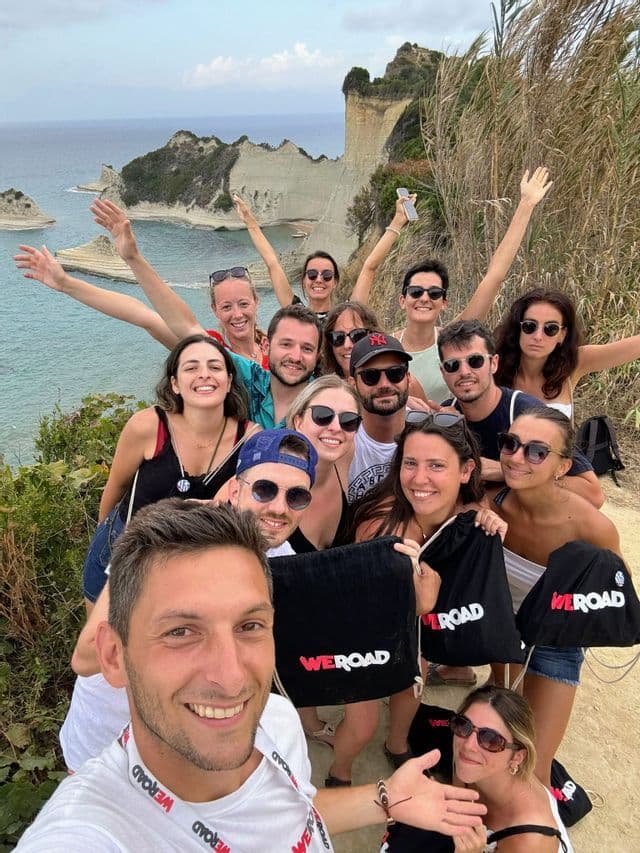 Un groupe WeRoad en voyage pose pour un selfie sur un sentier côtier panoramique, avec des formations rocheuses blanches et la mer en arrière-plan.