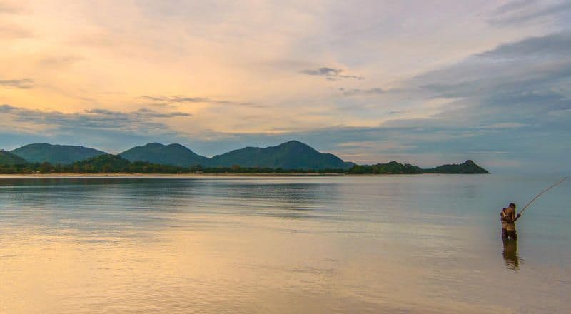 Una persona pesca con una canna nell'acqua calma fino alla vita, al tramonto, con montagne sullo sfondo.