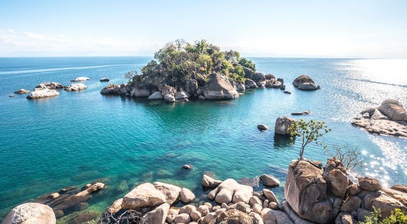 Un'isola rocciosa e alberata si trova in acque limpide e turchesi, circondata da grandi massi sotto un cielo soleggiato.