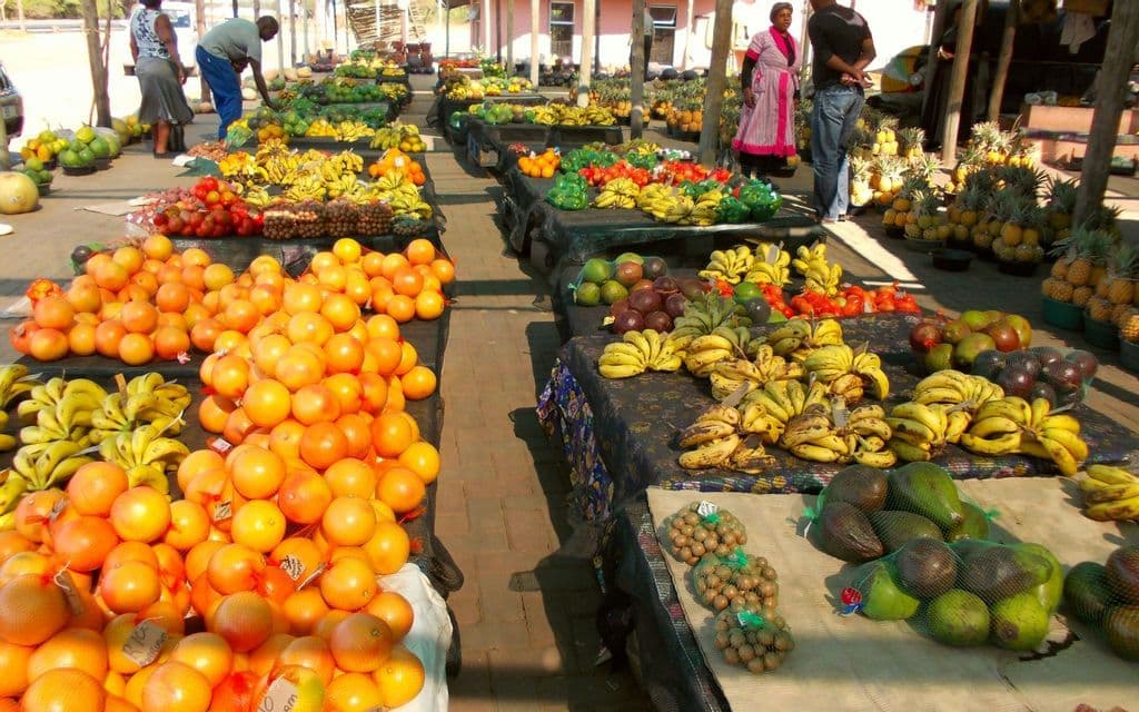 Un mercato della frutta all'aperto con lunghe bancarelle che espongono una varietà di prodotti freschi, tra cui arance, banane e avocado.