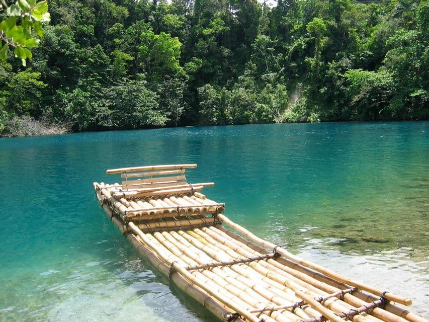 Ein Bambusfloß mit einer kleinen Bank treibt auf klarem, türkisfarbenem Wasser an einem Ufer, das von dichtem, grünem Wald gesäumt ist.