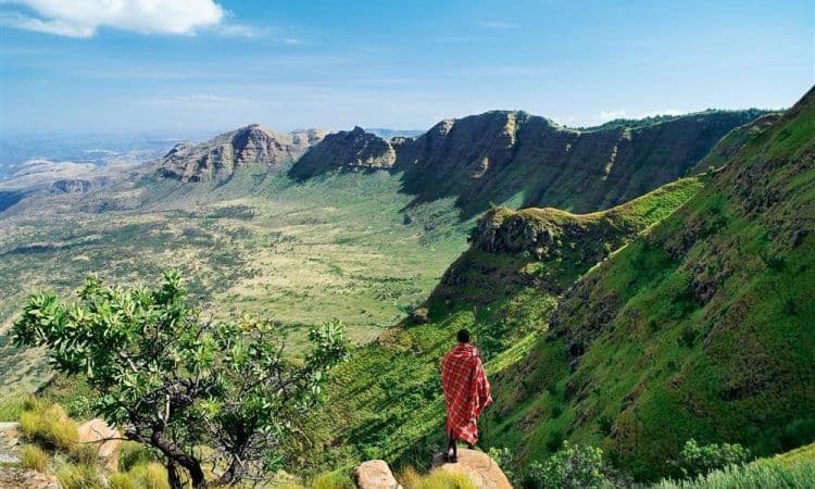 Una persona avvolta in una coperta rossa a fantasia si erge su una roccia, ammirando una vasta valle verde e una catena montuosa sotto un cielo blu.