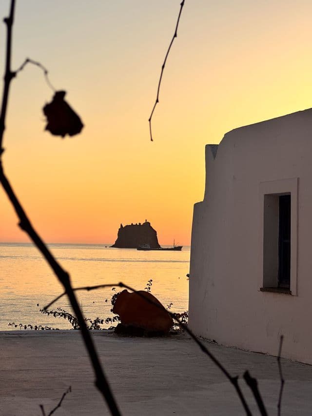 Una vista da una terrazza costiera mostra un isolotto roccioso sull'acqua stagliato contro un cielo arancione al tramonto, vicino a un edificio bianco.