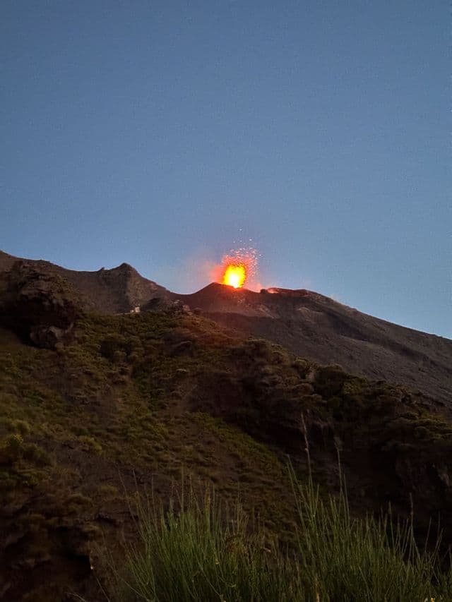 Un vulcano erutta al tramonto, scagliando uno spruzzo infuocato di lava e scintille nel cielo limpido dal suo cratere su un fianco della montagna.