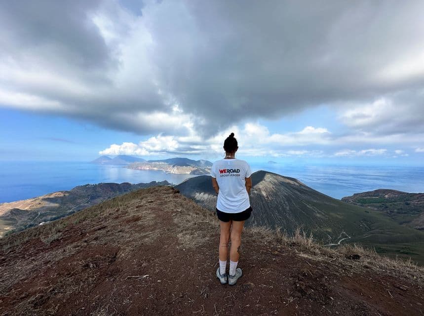 Un leader di gruppo WeRoad si trova su una cresta di montagna, con lo sguardo rivolto verso un cono vulcanico e una serie di isole nel mare.